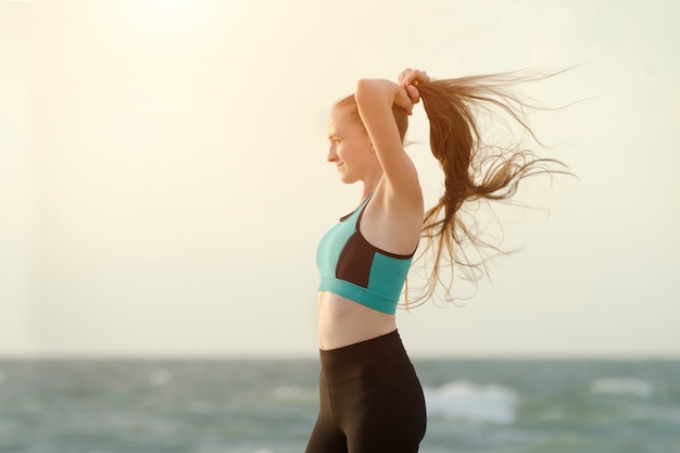Girl in sports clothes on the beach adjusts her hair. Morning light