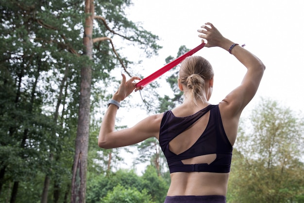 Girl in sports top and leggings trains with red elastic rope on sports grounds in the park