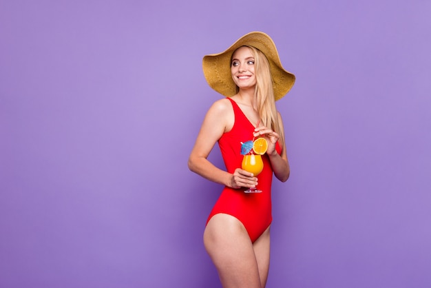 Girl standing half turned in hat and red swimsuit looking side holding cold summer cocktail