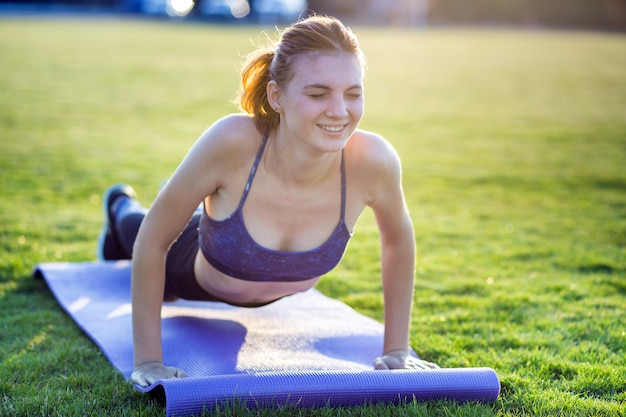 Girl standing in plank position with pain expression on grass in a city park