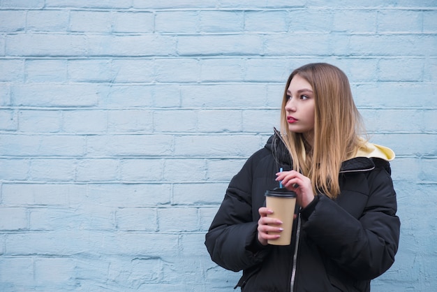 Girl stands on the background of a blue wall with a glass of coffee in her hands and looks to the side.