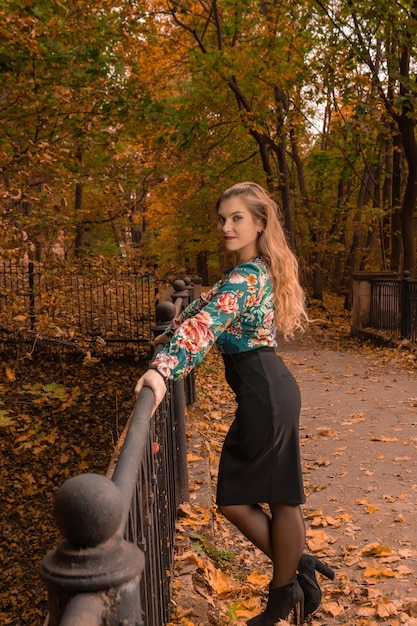A girl stands a bridge leaning on her side in the autumn woods in a coloured shirt and black skirt