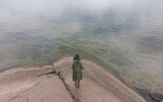 The girl stands on the granite shore of a clean lake Top view from behind