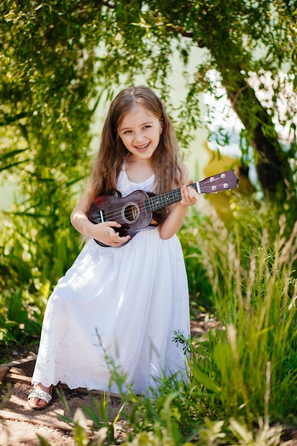 The girl stands on the grass, wears headphones and is learning\
to play ukulele strings, is learning outside the school in the\
nature park