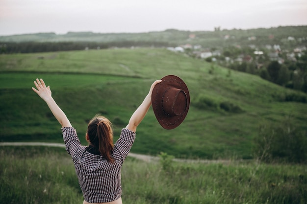 La ragazza sta in un campo verde con le braccia tese in diverse direzioni tenendo un cappello da cowboy