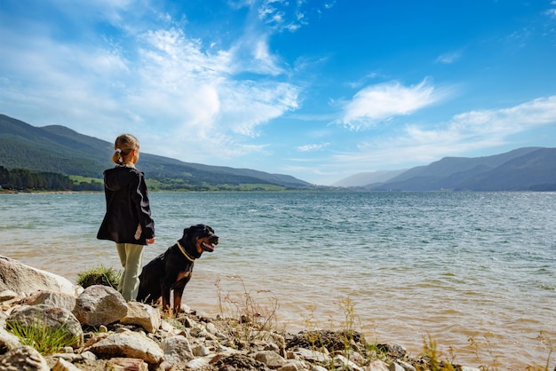 Girl stands near dog of Rottweiler breed on shore near lake against backdrop of mountain range covered with forest