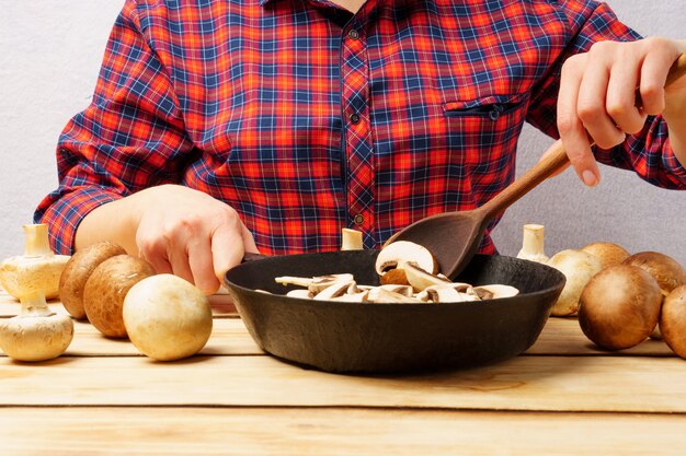 Девушка деревянной ложкой помешивает грибы на сковороде. Девушка в красной клетчатой рубашке готовит грибы на деревянном фоне.