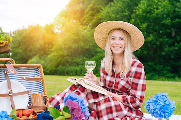 Girl in straw hat holding a wine glass and a book sitting in a picnic outdoors