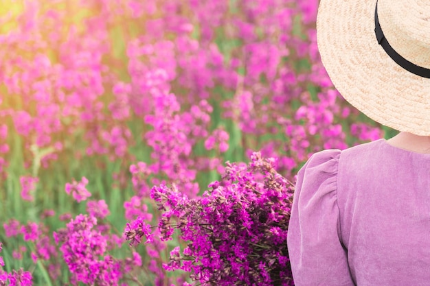 A girl in a straw hat and a purple dress enjoys the sunset with\
a bouquet of purple flowers in her hands copyspace