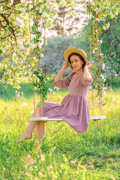 A girl in a straw hat and a purple dress poses on a rope swing in a blooming apple orchard