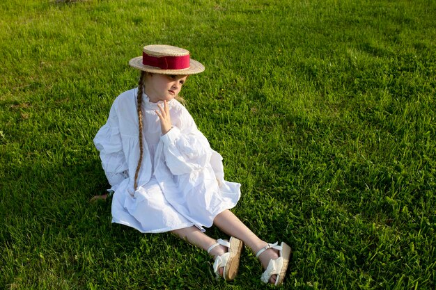 A girl in a straw hat sits on the grass and looks thoughtfully