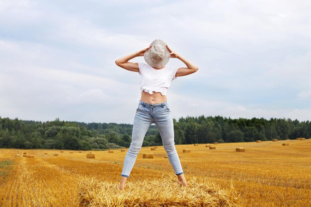 Girl in straw hat stands on a haystack on a bale in the agricultural field after harvesting