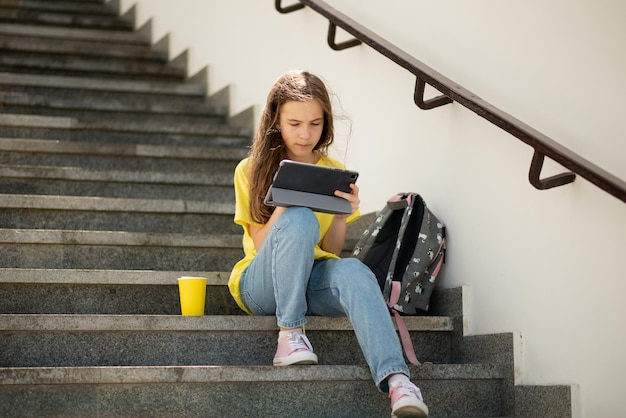 A girl on the street communicate on the gadget online. she sits\
on the steps. it\'s summer outside. video chat. communication\
online. technology. generation z.