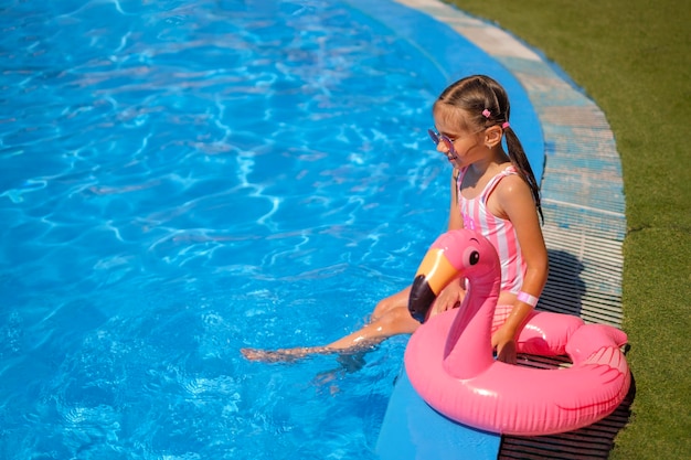 Girl in striped pink swimsuit is sitting on edge of pool