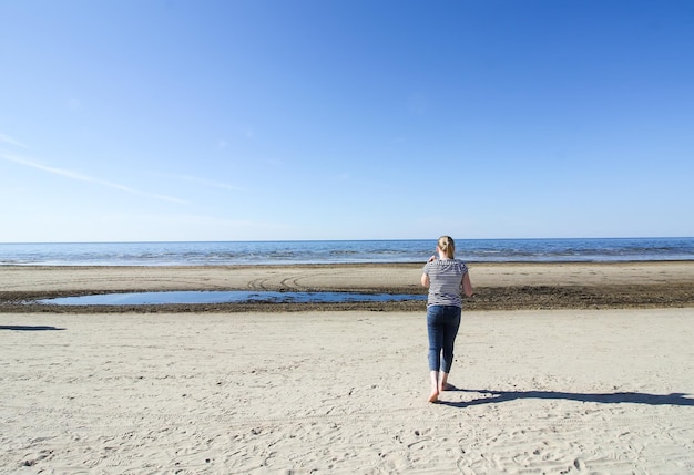 Girl in striped short and blue jeans walking on Baltic sea coast in sunlight