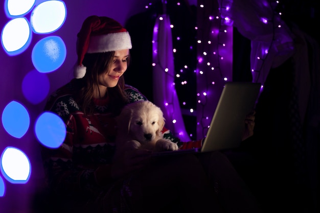 クリスマスの服の女子生徒は子犬とラップトップで夜家に座っています。