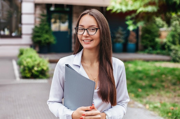 A girl student in glasses holds a folder and smiles against the background of the building
