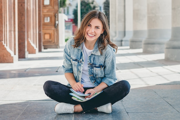 Girl student sits opposite the university and smiling. Cute girl student holds pensil, folders, notebooks and laughs. Girl teaches lessons