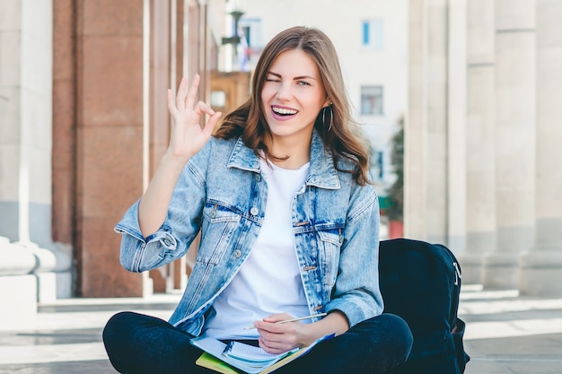 Photo girl student sitting near the university and shows okay gesture with his hands