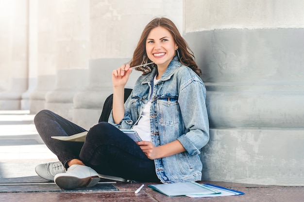 Foto studentessa che si siede vicino all'università e sorridere. studentessa carina detiene cartelle, quaderni e risate. la ragazza insegna lezioni