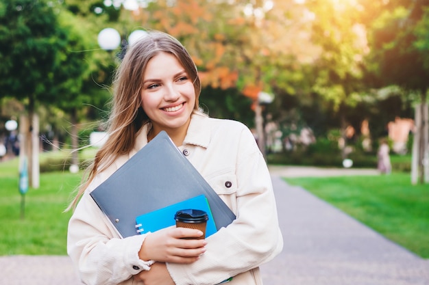 Girl student smiles and walks in the park with notebooks and a cup of coffee. 