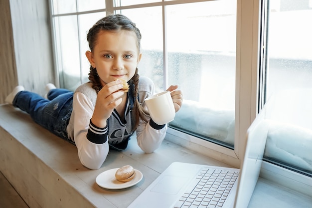 La studentessa studia online con l'insegnante di videochiamata, la ragazza felice ascolta la lezione guarda al computer portatile a casa e beve il tè e? mangiare i biscotti. covid-19-coronavirus.