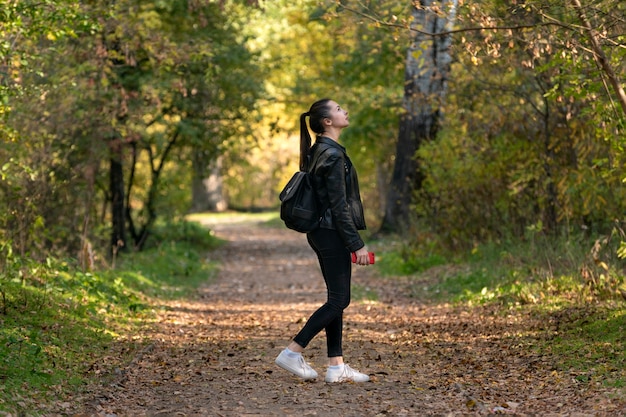 女子学生が秋の公園を散歩します。スタイリッシュな若い女性が森の中を歩きます。黄色い落ち葉のある路地。