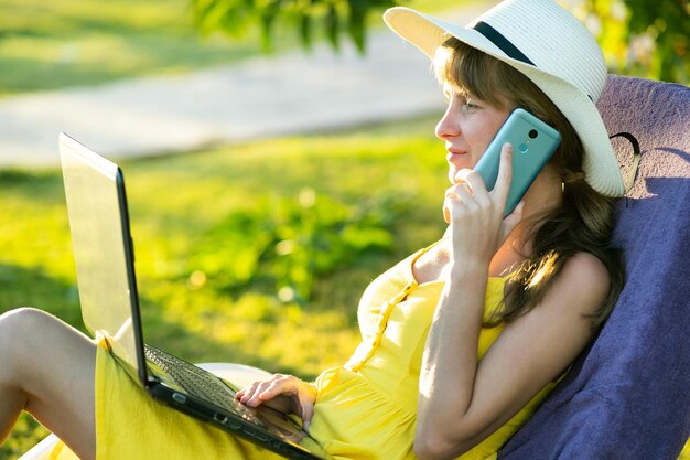 Foto studentessa in abito estivo giallo che riposa sul prato verde nel parco estivo che studia sul computer portatile con conversazione sul cellulare facendo affari e imparando durante il concetto di quarantena