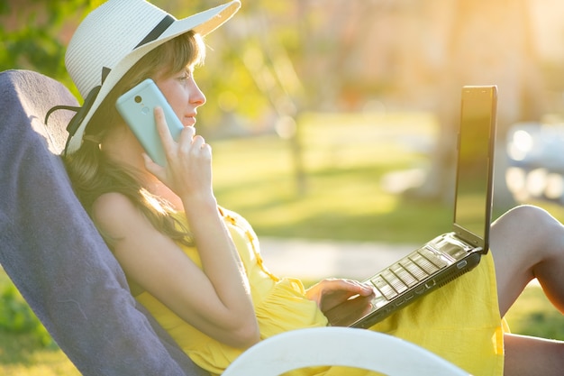 モバイル携帯電話で会話しているコンピューターのラップトップで勉強して夏の公園の緑の芝生で休んでいる黄色の夏のドレスの女子生徒。検疫の概念の間にビジネスを行い、学習します。