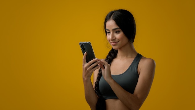 Photo girl in studio yellow background indian 20s sporty woman in sportswear with smartphone scrolling