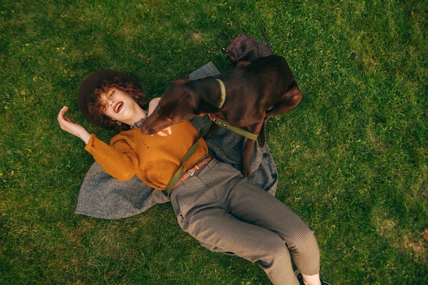 Girl in stylish clothes lying on green grass and playing with brown young dog