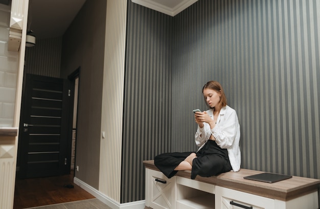 Girl in stylish clothes sits at home on a bedside table in the apartment and uses a smartphone