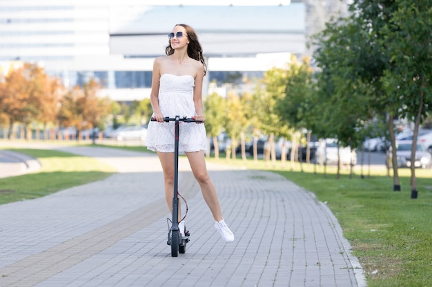 A girl in a summer dress rides an electric scooter on the sidewalk