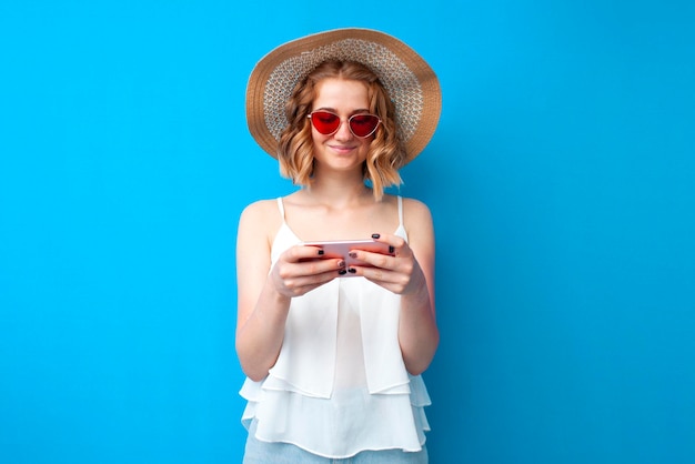 Girl in a sun hat and glasses use a smartphone on a blue isolated background