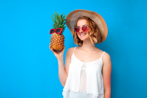 Girl in a sun hat and glasses with pineapple smiles and looks at the copyspace