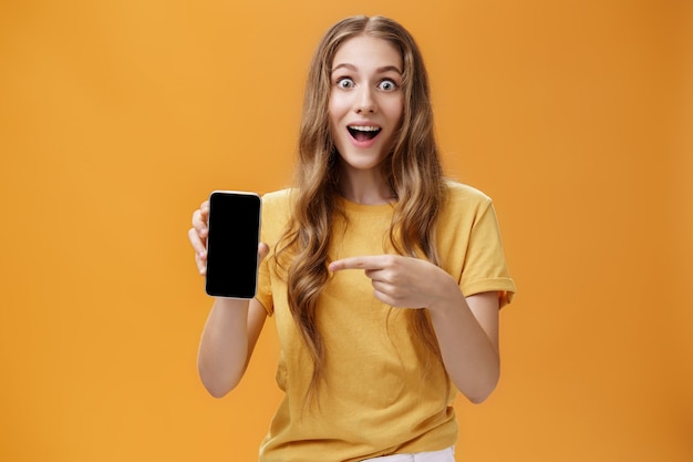 Ragazza sorpresa dalle fantastiche funzioni dello smartphone. ritratto di donna snella europea sveglia felice e stupita con acconciatura naturale ondulata che mostra il cellulare che punta allo schermo del gadget all'app. copia spazio