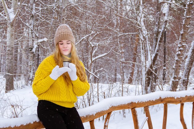 Girl in a sweater holding a cup thermos cap with a hot drink in a winter park