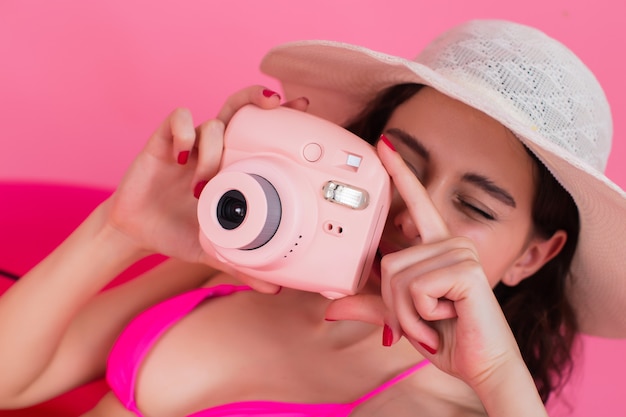 The girl in a swimsuit lies on an inflatable flamingo and takes a photo on the instant camera