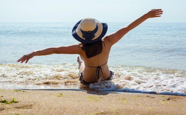 ビーチで水着と麦わら帽子をかぶった女の子は、晴れた夏の日に海岸で波に座って後ろ姿の女性に到達しました