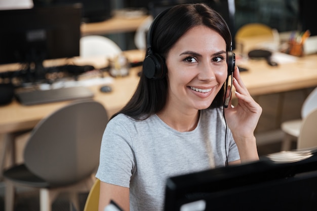 Girl in t-shirt sitting by the computer in headphone and looking at camera. call center.