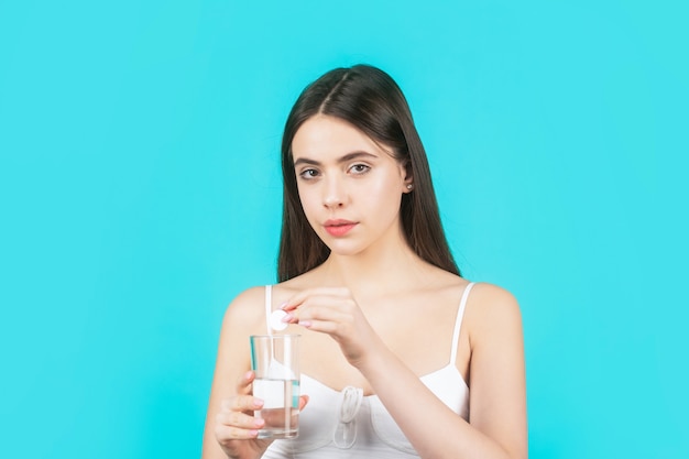 La ragazza prende alcune pillole, tiene un bicchiere d'acqua, isolato sull'azzurro. giovane donna che prende pillola contro il mal di testa. bruna che prende una pillola con un bicchiere d'acqua. donna che assume farmaci per alleviare il mal di testa.