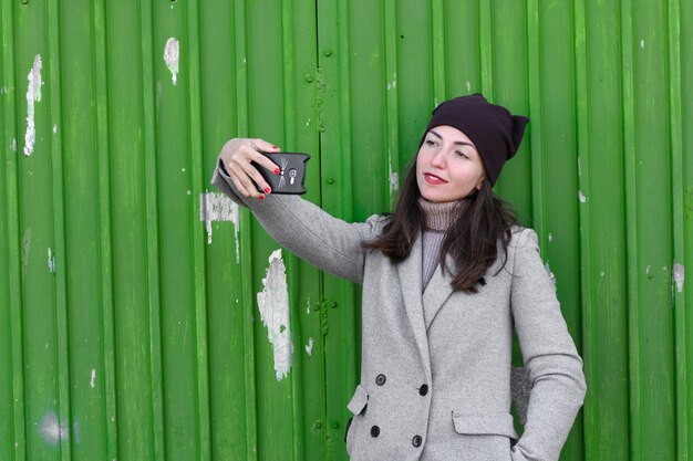 여자는 녹색 산업 문에는 selfie를 걸립니다. 모자와 코트를 입고. 쓰기위한 장소. 차가운 탑을 입고 있습니다. 옷과 스타일. 자연스러운 감정