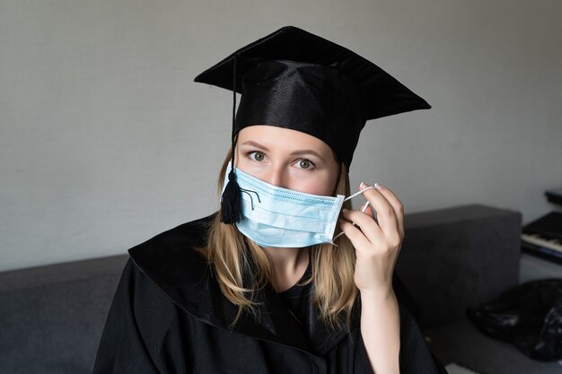 Girl taking off his medical mask with graduation hat on grey background