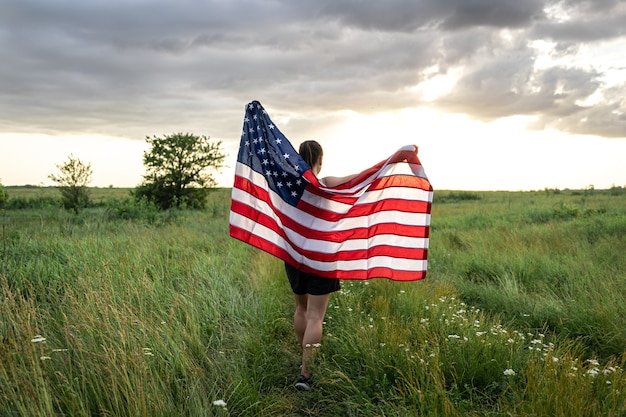 Girl teenager female young woman in a field wrapped in USA stars and stripes flag in evening sunshine.