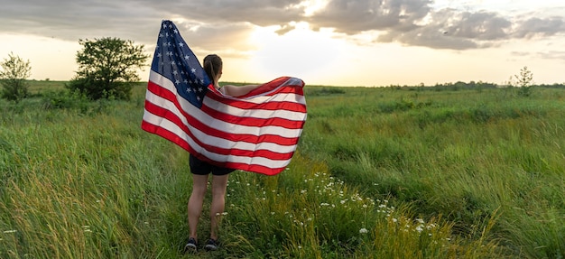 Girl teenager female young woman in a field wrapped in usa stars and stripes flag in evening sunshine.