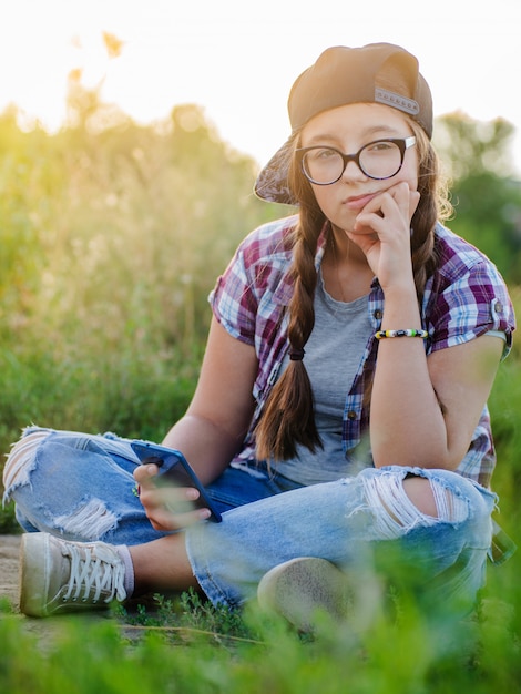 メガネの10代の少女は、selfieスマートフォンのヘッドフォンで地上都市に座っています。
