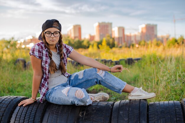 メガネの10代の少女は、selfieスマートフォンのヘッドフォンで地上都市に座っています。
