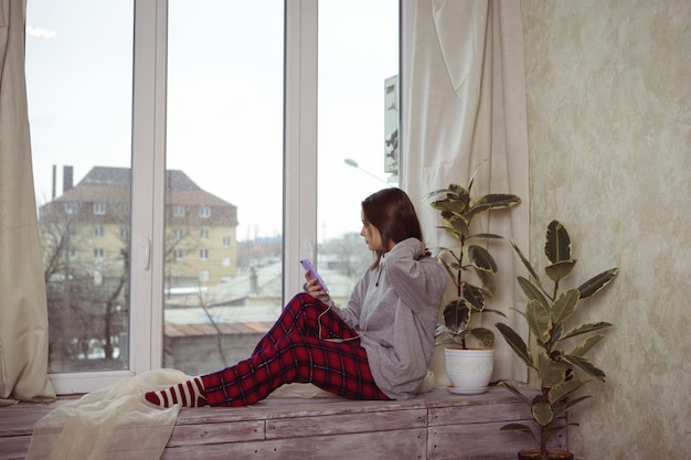 Фото Девочка-подросток сидит на окне с телефоном в руке и слушает что-то в наушниках. одиночество. молодая женщина смотрит в окно