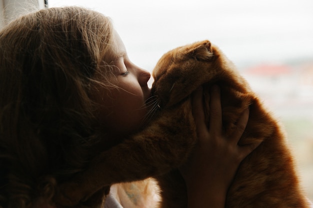 Foto l'adolescente si siede in pigiama alla finestra della casa e tiene in mano un gatto allo zenzero. stare a casa.