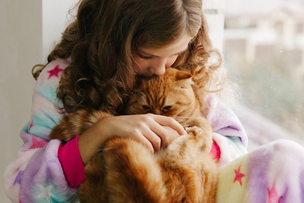 Girl teenager sits in pajamas at the window of the house and holds a ginger cat. stay home.